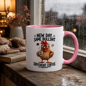 Può includere: Una tazza in ceramica bianca con interno e manico rosa, raffigurante un pollo marrone imbronciato che tiene in mano una piccola tazza di caffè. Il testo recita: NEW DAY, SAME BULLSHIT, DIFFERENT COFFEE. Il pollo ha piume rosse sulla testa.