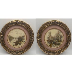 May include: Two antique framed prints. Each print features a detailed landscape scene within a pink mat, encased in a gold-colored, ornate, round frame. The prints depict historical scenes with figures and buildings.