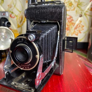 May include: A vintage black Kodak Vigilant Junior camera with a Kodet lens and Dak Shutter. The camera features a collapsible bellows design and is set on a red surface. The lens has the following settings: 1/12.5 16 22 32.