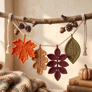 Op de afbeelding: Een decoratieve slinger met vier gehaakte bladeren in oranje, beige, bordeauxrood en olijfgroen, opgehangen aan een natuurlijke houten tak met een dik crèmekleurig touw, versierd met kleine bruine dennenappels en eikels.