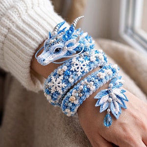 May include: A crochet dragon bracelet in shades of blue and white, featuring intricate needlework, silver-toned horns, white snowflake embellishments, small pearl-like beads, and a dangling blue teardrop crystal charm at the end.