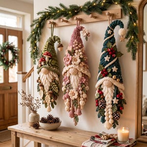 May include: Three hanging gnome ornaments crafted from yarn, featuring pointed hats in olive green, dusty rose, and dark teal. Each gnome has a cream-coloured braided beard, decorated with various embellishments like felt mushrooms, fabric flowers, wooden beads, pinecones, and small stars.