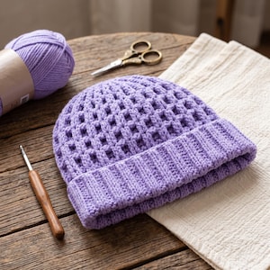May include: A lavender crochet beanie featuring a waffle-like grid pattern on the crown and a ribbed brim. The beanie is accompanied by a matching ball of lavender yarn, a metal crochet hook with a wooden handle, and a pair of small scissors.