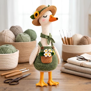 May include: A crocheted goose doll wearing a straw hat with a sunflower, a green neckerchief, and green overalls with a small basket of white flowers attached to the front. The goose has a cream-colored body, orange beak, and orange webbed feet.