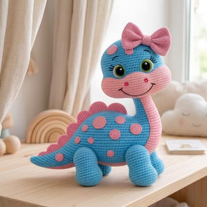 May include: A crocheted dinosaur plush toy featuring a bright blue body with pink spots, a pink snout, and a pink bow on its head. The creature has large green eyes with long eyelashes and a stitched smile, with a row of pink spikes along its back.