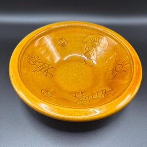Op de afbeelding: Een ronde, glanzende mosterdgele keramische kom met donkerbruine, gegraveerde lijntekeningen op het binnenoppervlak. De gravures tonen gestileerde figuren, een zon, een spiraal en een cirkelvormig patroon rondom de kom.