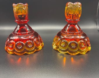 Puede incluir: Un par de portavelas de vidrio ámbar y rojo con un patrón prensado de lunas y estrellas. Cada portavelas tiene una base ancha festoneada y una sección superior cilíndrica diseñada para sostener una vela de forma segura.