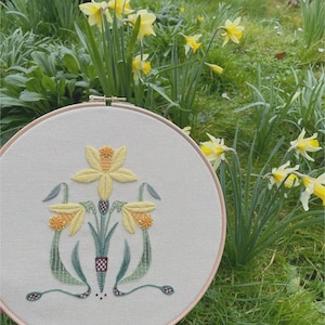 May include: A circular wooden embroidery hoop containing a light gray fabric panel with a hand-stitched design of three yellow daffodils. The embroidery features green stems and leaves with intricate cross-stitch details and orange centers.
