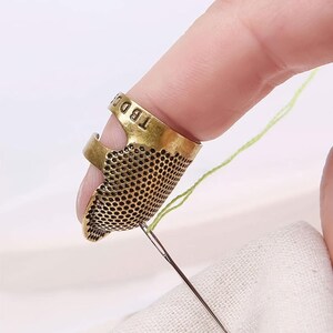 May include: An antique-style brass open-sided thimble featuring a textured honeycomb pattern on the exterior and the letters TBDD embossed on the band. The thimble is worn on a finger while a silver sewing needle pulls a light green thread.