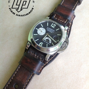 Könnte beinhalten: Ein braunes Lederarmband mit weißen Nähten. Das Armband ist an einer silbernen Uhr mit schwarzem Zifferblatt und weißen Zeigern befestigt. Das Zifferblatt der Uhr trägt den Text "Luminor Panerai Automatic" und "Power Reserve".