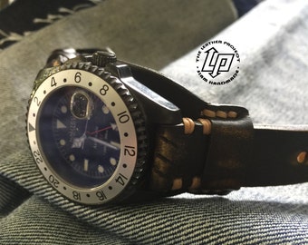 May include: A black and silver watch with a blue face and a brown leather strap. The strap has a distressed look and is stitched with a light brown thread. The watch face has a white bezel with black numbers and markers. The watch is on a denim background.