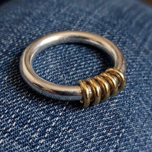 May include: A circular ring featuring a smooth, polished silver-toned band. A section of the band is adorned with five small, interconnected gold-toned rings that wrap around the main silver structure, creating a contrasting metallic layered design.
