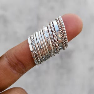 Può includere: Una collezione di dieci anelli color argento impilati insieme, caratterizzati da varie texture tra cui fasce lisce, disegni a corda intrecciata e incisioni complesse con motivi geometrici e floreali, tutti disposti in un'unica fila.
