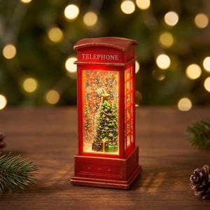 Könnte beinhalten: Eine rote, rechteckige, beleuchtete Schneekugel in Form einer Telefonzelle mit einem geschmückten Weihnachtsbaum im Inneren. Das Wort TELEPHONE steht in Weiß auf dem oberen Paneel. Das Innere ist mit wirbelndem Glitzer gefüllt.