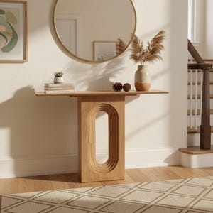 Peut inclure: Une console en chêne clair dotée d'un pied central avec une découpe ovale creuse et étagée. La table présente un plateau rectangulaire plat et est placée contre un mur sous un grand miroir circulaire avec un cadre fin de couleur dorée.