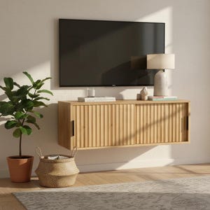 Könnte beinhalten: Ein wandmontiertes Medienboard aus hellem Holz mit zwei Schiebetüren in vertikaler Rillenoptik und schwarzen Griffen. Das Board ist unter einem Flachbildfernseher angebracht und trägt eine Tischleuchte sowie einige Bücher.