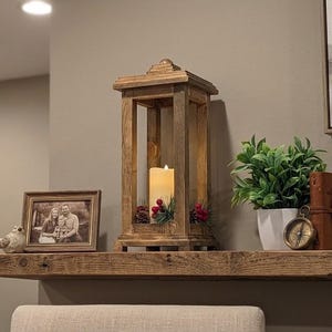 May include: A tall, rectangular wooden lantern with a natural finish, containing a white pillar candle surrounded by small pinecones and red berries. The lantern sits on a wooden shelf alongside a framed photograph, a small bird figurine, a potted green plant, a brass compass, and a brown leather-bound journal.
