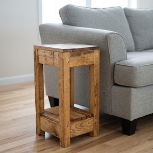 May include: A tall, rectangular side table constructed from light brown wood with a visible grain pattern. The table features four sturdy legs, a flat square top surface, and a lower shelf positioned between the legs near the base of the unit.