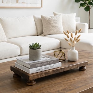 May include: A rectangular dark brown wooden riser with four rounded feet, holding two stacked books titled Modern Living and White Interiors, a small green succulent in a gray pot, a gold geometric wire sculpture, and a white vase with dried grass.