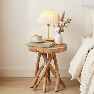 May include: A round side table crafted from natural light-toned wood with four interlocking branch-like legs. The table surface holds a pleated white lamp, a ceramic teacup on a saucer, a green book, and a white vase with dried floral stems.