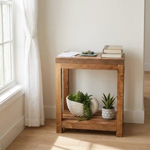 May include: A rectangular wooden console table with a natural brown finish, featuring a flat top surface and a lower shelf. The top holds two stacked books and a small tray with keys. The lower shelf displays a white shell-shaped planter with green succulents and a small gray patterned pot with a green plant.