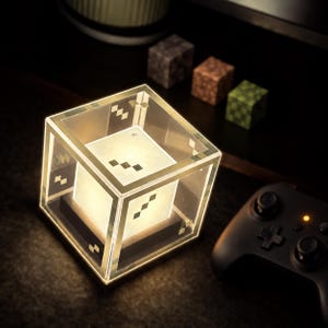 Peut inclure: Une lampe carrée lumineuse dotée d'un cadre extérieur transparent avec des accents blancs pixélisés et un cube intérieur émettant une lumière douce. Le design présente une esthétique en blocs avec une base noire et une structure lisse.