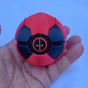 May include: A spherical object featuring a textured red surface with black accents and a central circular emblem displaying the white eyes and red outline of the Deadpool character logo. The item has a segmented design with a matte finish.