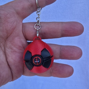 May include: A spherical keyring featuring a textured red surface with black geometric accents and a central circular emblem displaying a black and red Deadpool-inspired logo, attached to a silver-toned metal split ring and chain.