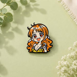 May include: A metal enamel pin featuring a stylised anime character with long orange hair, large red eyes, and a wide smile. The character wears a yellow and black patterned top and has a blue tattoo on the upper arm, outlined in black metal.