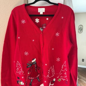 Könnte beinhalten: Eine leuchtend rote Langarm-Strickjacke mit Knopfleiste, verziert mit gestickten weißen Schneeflocken und einer Winterszene am Saum. Das Motiv zeigt eine Person in rotem Mantel und Schal, die einen Schlitten mit Hund zieht, sowie einen weiteren Hund.
