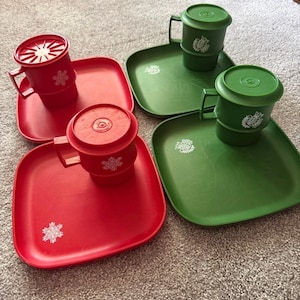 May include: A set of four square plastic plates and four matching mugs with lids. Two plates and two mugs are red with white snowflake designs, while two plates and two mugs are green with white floral patterns on the mugs and plates.