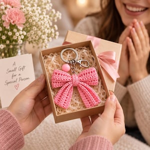 Può includere: Un portachiavi a forma di fiocco lavorato all'uncinetto rosa con un anello in metallo argentato, un moschettone e una piccola perlina rosa, presentato in una scatola regalo in cartone riempita di carta, accompagnato da un biglietto con il testo A Small Gift for a Special Person.