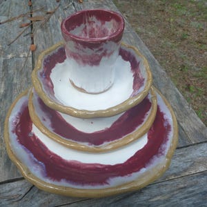 May include: A ceramic dinnerware set consisting of a cup, a small bowl, and a large plate stacked vertically. Each piece features a white center, a deep maroon glazed ring, and a thick, textured tan rim with an irregular, hand-formed edge.