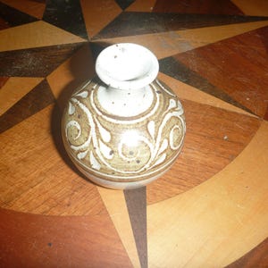 May include: A small, round ceramic vase with a narrow neck and a flared rim. The vase features a cream-colored base with brown, hand-painted swirling leaf patterns and dark brown speckles distributed across the surface of the pottery.