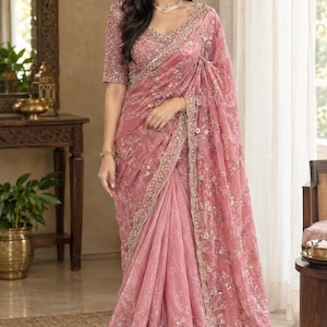 May include: A dusty rose saree featuring intricate gold embroidery, sequins, and floral patterns throughout the fabric. The garment includes a matching short-sleeved blouse with a V-neckline, heavily embellished with gold thread and beadwork.