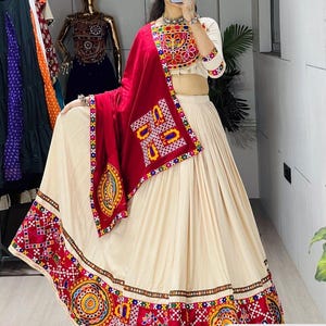 May include: A cream-colored lehenga choli set featuring a cropped blouse and a long, pleated skirt. The garment is adorned with vibrant, multi-colored embroidered patterns in red, yellow, and blue, including a matching red dupatta with embroidery.