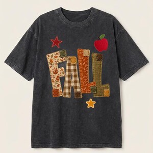 May include: A charcoal gray short-sleeve t-shirt featuring the word FALL in large, fabric-appliqued letters with various brown, orange, and green patterns. The design includes a red apple, a red star, and a yellow star stitched onto the front.