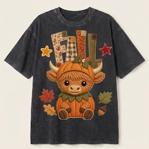 Peut inclure: Un t-shirt gris anthracite délavé à l'acide avec un motif représentant une vache Highland assise dans une citrouille. Au-dessus de la vache, le mot FALL est écrit en lettres à motifs. Le design inclut des feuilles d'automne, un gland et deux étoiles.