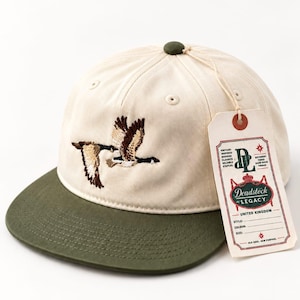 May include: A cream-coloured baseball cap with an olive green brim and a dark green button on top. The front features an embroidered design of two flying geese. A rectangular paper tag is attached, displaying the text Deadstock LEGACY.