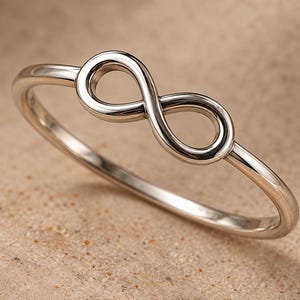 Op de afbeelding: Een gepolijste zilverkleurige metalen ring met een doorlopend oneindigheidssymbool in het midden van een dunne, gladde band. De ring heeft een reflecterende, metalen afwerking en een eenvoudige, strakke constructie zonder extra stenen.