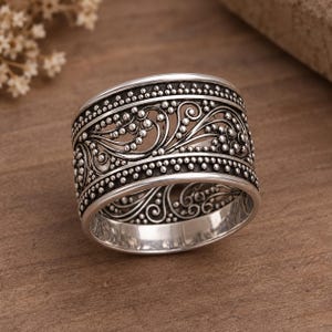 Puede incluir: Un anillo de banda ancha de plata con un intrincado diseño calado de patrones de filigrana en espiral. La superficie está acentuada con pequeñas cuentas de plata en relieve a lo largo de los bordes superior e inferior, creando un acabado texturizado y oxidado.