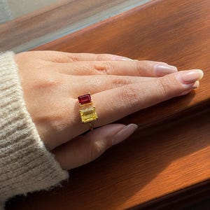 Puede incluir: Un anillo abierto de oro con dos piedras preciosas de corte esmeralda. Una piedra es de color rojo intenso y la otra es de color amarillo brillante. El anillo está colocado en un dedo, mostrando las dos piedras rectangulares en la banda.