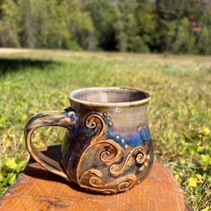 May include: A ceramic mug with a rounded body and a curved handle. The exterior features a glossy glaze in shades of deep blue, brown, and tan, decorated with raised, swirling scroll patterns and small dots in a lighter, warm beige tone.