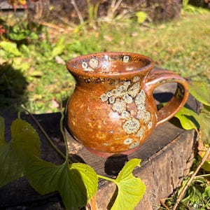 May include: A round ceramic mug with a glossy, mottled brown and amber glaze featuring distinct, light-colored crystalline patterns across the surface. The mug has a wide, flared rim and a sturdy, curved handle attached to the side.