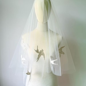 May include: A white tulle wedding veil featuring delicate, embroidered swallow bird motifs scattered across the sheer fabric. The veil is draped over a mannequin, showcasing the intricate bird patterns in a soft, light-colored thread design.