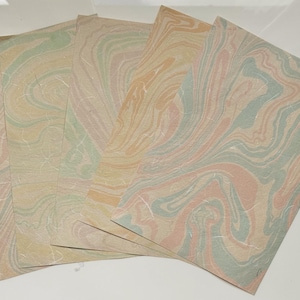Può includere: Un set di cinque fogli di carta rettangolari caratterizzati da un motivo marmorizzato con colori vorticosi di verde salvia, pesca tenue e crema. La carta presenta una texture fibrosa visibile, creando un effetto a strati sui fogli sovrapposti.