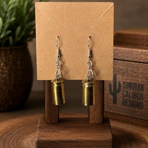 May include: A pair of drop earrings featuring polished brass bullet casings suspended from silver-toned metal fishhook wires. The earrings are displayed on a rectangular brown card, which is mounted on a small wooden stand for presentation.