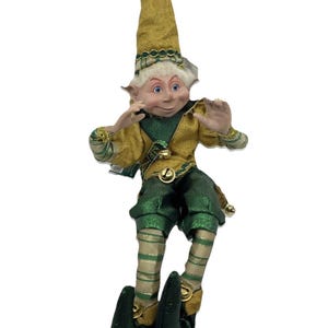 May include: A decorative elf figurine wearing a gold and green outfit with a matching pointed hat. The elf has white hair, pointed ears, and blue eyes. The clothing features gold bells on the hat, collar, and shoes. The legs are striped in gold and green, and the shoes are dark green.