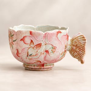 May include: A ceramic cup with a scalloped rim and a handle shaped like a fan. The exterior features a hand-painted design of a sleeping fox surrounded by pink, cloud-like patterns and small floral accents in shades of pink, white, and green.