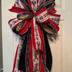 May include: A large decorative bow featuring a festive nutcracker pattern on a cream background, accented with solid red velvet, black velvet, and glittery red, green, and blue striped ribbons. The bow has multiple long, flowing tails.